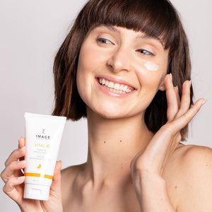 Smiling woman with short brown hair applies VITAL C hydrating enzyme masque | IMAGE Skincare to her cheek, holding the skincare product tube against a plain background.