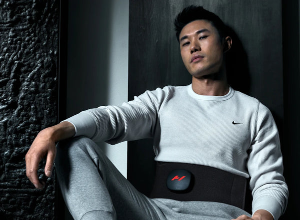 Man wearing a Venom 2 Back | Hyperice recovery device around his lower back, sitting relaxed against a dark wall in casual athletic wear, showcasing wellness technology for body care and recovery.