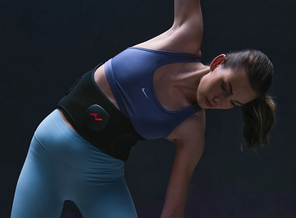 Woman wearing a Venom 2 Back | Hyperice recovery device on her lower back while stretching in athletic wear, promoting wellness and body care.