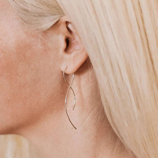 Close-up of a woman wearing delicate, slender silver earrings with a flowing twisted design, showcasing the elegance of Vista Earrings | Purpose Jewelry.
