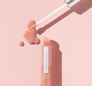 An open tube of Vitamin Glaze® Oil Infused Lip Gloss – Peach Peony | Farmhouse Fresh with a wand applicator hovering above a glossy peach-colored spill on a soft pink background.