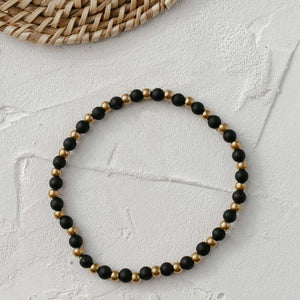 Vivid Bracelet | Purpose Jewelry featuring alternating black and gold round beads on a stretch band, styled on a textured white surface with natural woven decor.