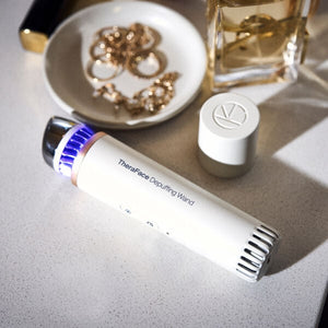 TheraFace Depuffing Wand | Therabody lying on a countertop next to a small dish with gold jewelry and a glass perfume bottle, highlighting a sleek skincare wellness device.