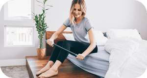 A smiling woman using a massage roller on her legs while sitting on a bed in a bright, cozy room featuring the RAD All In Kit | RAD wellness recovery tool.