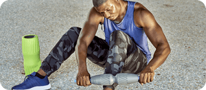 Man using a gray handheld recovery tool on his leg outdoors, wearing a blue tank top and camo leggings, next to a green foam roller, showcasing the RAD All In Kit | RAD for wellness and body care.