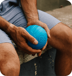 Person holding a blue RAD ATOM | RAD massage ball designed for muscle recovery and body care, sitting outdoors in casual athletic wear.