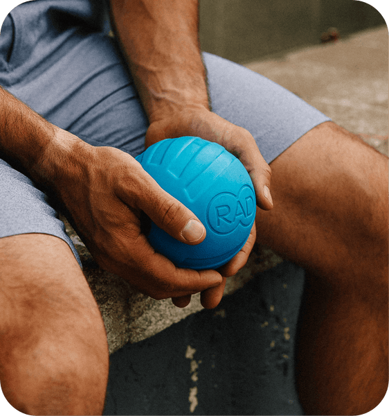Person holding a blue RAD ATOM | RAD massage ball designed for muscle recovery and body care, sitting outdoors in casual athletic wear.