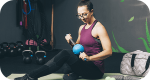 Woman in a gym setting using a blue RAD ATOM | RAD massage and recovery tool on her arm, surrounded by kettlebells and workout accessories.
