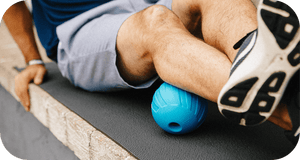 Person using a blue massage roller under their calf while seated outdoors on a mat, demonstrating recovery and wellness with the RAD ATOM | RAD product.