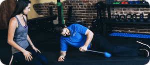 Man using RAD ATOM | RAD blue massage tool on side while a woman watches in gym setting, demonstrating recovery and wellness device for body care and muscle relief.