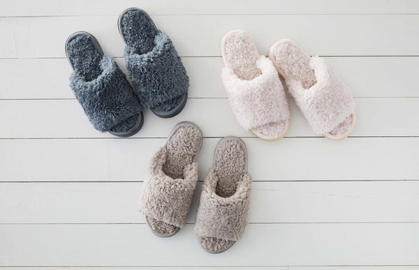 Three pairs of plush open-toe slippers in dark gray, beige, and ivory shades arranged on a white wooden floor, highlighting cozy textures and comfort with the Anywhere Berber Slide | Lemon.