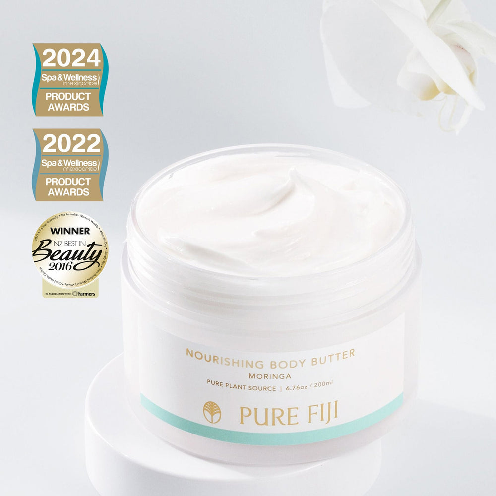 Open jar of Nourishing Body Butter | Pure Fiji with creamy white texture, placed on a white surface with a white flower and spa and wellness product award badges in the background.