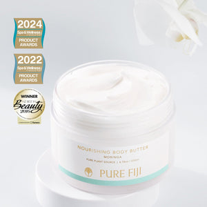 Open jar of Nourishing Body Butter | Pure Fiji with creamy white texture, placed on a white surface with a white flower and spa and wellness product award badges in the background.