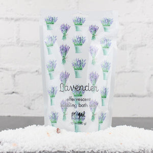 White resealable pouch with illustrations of lavender bouquets containing Lavender Bubbling Bath Salts | Primal Elements surrounded by coarse white bath salts on a gray surface against a white brick background
