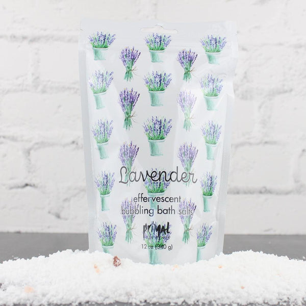 White resealable pouch with illustrations of lavender bouquets containing Lavender Bubbling Bath Salts | Primal Elements surrounded by coarse white bath salts on a gray surface against a white brick background