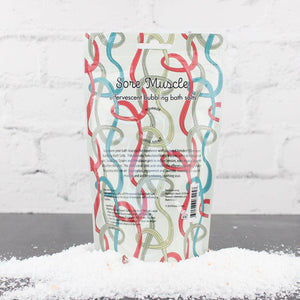 Package of Sore Muscle Bubbling Bath Salts | Primal Elements standing upright on a gray surface with colorful abstract line patterns on the packaging and scattered bath salts around the base.