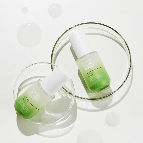 Two green and white dropper bottles of Calming Biotics Blemish Serum | Leaders placed on clear glass petri dishes with drops of liquid on a white surface, highlighting skincare and wellness.