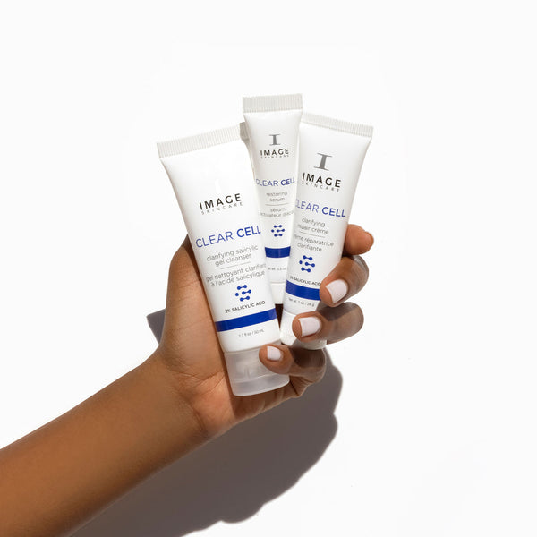 Hand holding CLEAR SKIN SOLUTIONS Blemish Defense Trio | IMAGE Skincare, featuring three skincare tubes from the Clear Cell line against a white background.