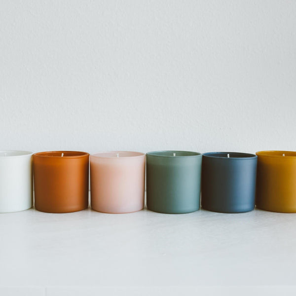 A row of six glass jar candles in varying muted colors including white, terracotta, blush pink, sage green, slate blue, and mustard, featuring the Evergreen + Eucalyptus Dignity Series Glass Jar Soy Candle | Calyan Wax Co. - 5.3 oz.