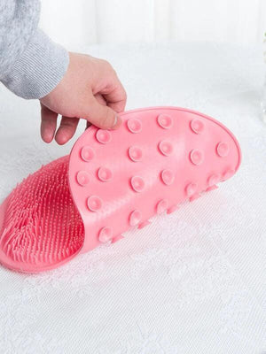 A hand holding a flexible pink silicone scrubber with textured bristles and suction cups, designed for skincare and body care use with the Back & Foot Scrubber | Dreambox Beauty.