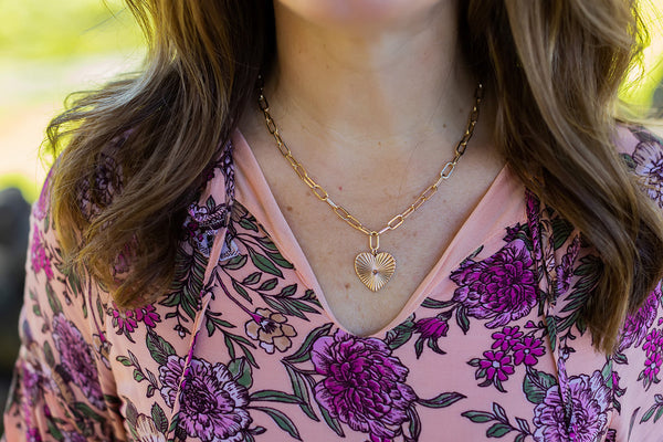 Close-up of a woman wearing the Full Heart Necklace | Meghan Bo, featuring a gold heart pendant with a sunburst design on a delicate chain, styled with a floral pink blouse.