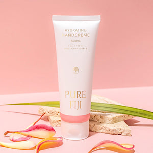 Hydrating Hand Crème | Pure Fiji in a white and pink tube with guava scent, displayed on a soft pink background with tropical leaves and petals for a fresh skincare vibe.