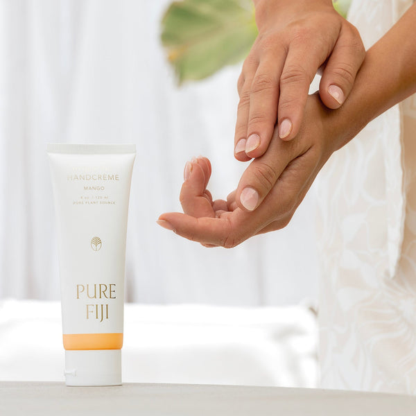 Close-up of hands gently applying Hydrating Hand Crème | Pure Fiji with a tube of mango-scented hand cream in soft natural light, emphasizing skincare and wellness.