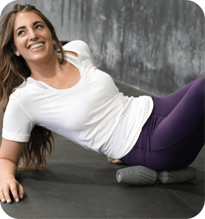 A woman using the RAD HELIX | RAD body recovery tool in a gym setting, rolling out her side muscles while wearing a white top and purple leggings.