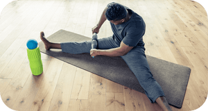 Man using a textured RAD HELIX | RAD massage roller on his leg while sitting on a yoga mat in a bright wooden floor room for muscle recovery and wellness.