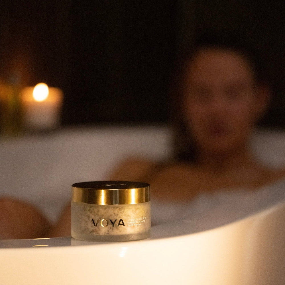 A container of Indulgent Sea Marine Bath Salts | VOYA placed on a bathtub edge with a softly lit candle in the background and a person relaxing in a warm bath, evoking a serene spa atmosphere.