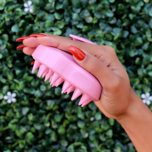 Hand holding a pink silicone scalp scrubber with soft bristles against a green leafy background, designed for gentle scalp massage and cleansing, Scalp Scrubber [Revitalizing Scalp Massager] | Dreambox Beauty.