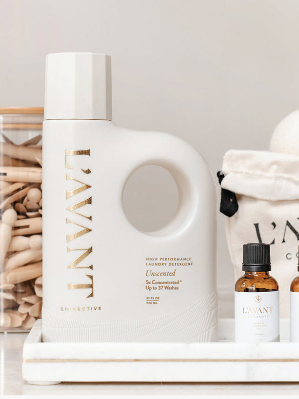 White bottle with a circular handle of High Performing Laundry Detergent - Unscented | L'AVANT Collective displayed on a white tray alongside small amber bottles and a fabric pouch.