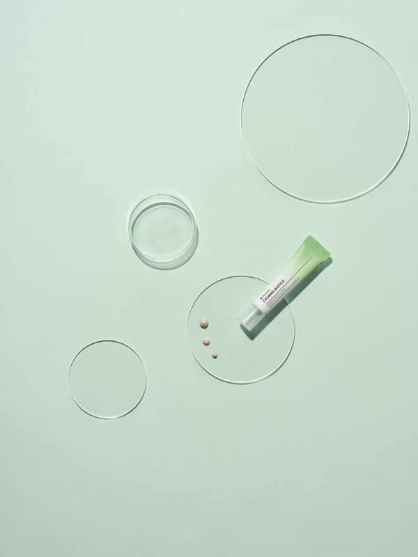 Tube of Calming Biotics Blemish Spot Cream | Leaders on a pale green surface with clear round glass plates and small cream dots, showcasing the skincare product for targeted blemish care.