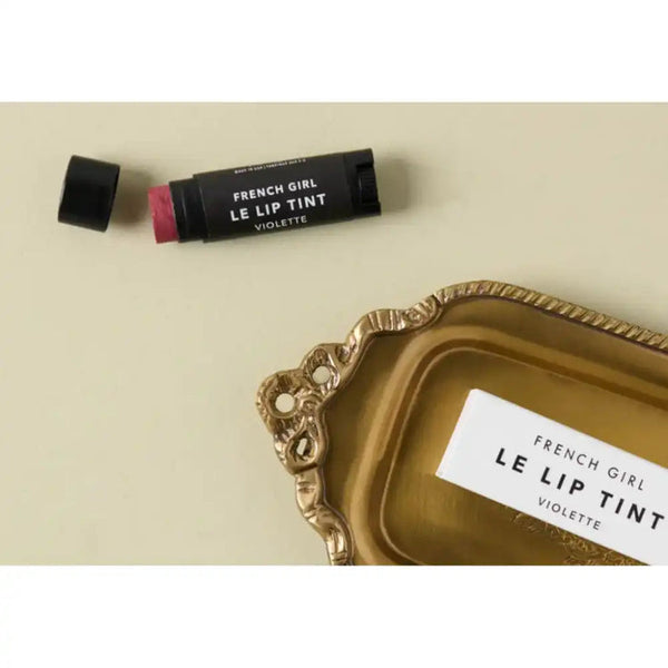Le Lip Tint | French Girl in a muted violet shade displayed next to a decorative gold tray, highlighting a chic and elegant lip care product.