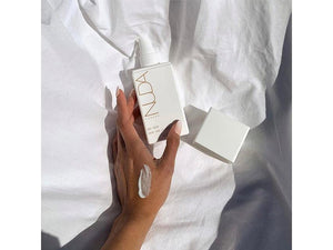Hand applying white Body Cream | NUDA from a sleek, minimalist pump bottle on a soft white fabric background, highlighting skincare and body care wellness.