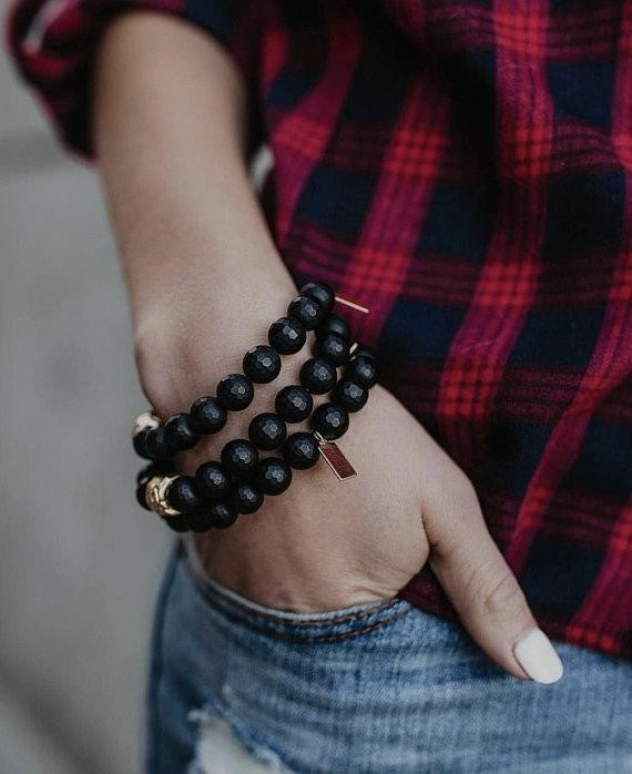 Close-up of a wrist wearing a Matte Black Beaded Stretch Bracelet | Meghan Bo, featuring multiple strands of polished black beads and gold accents against casual plaid and denim clothing.