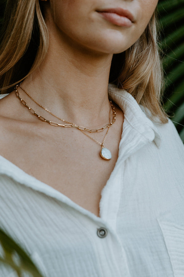Close-up of a woman wearing layered gold chains with a round white pearl pendant, showcasing the elegant Pearl Necklace | Meghan Bo against a soft white textured shirt.
