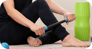 Person using the Rod Mini | RAD massage tool to relieve foot and leg muscle tension while sitting on the floor near a green foam roller in a wellness setting.