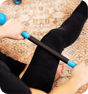 Person using a Rod Mini | RAD massage tool with blue handles on their leg while sitting on a patterned rug, promoting muscle recovery and wellness.