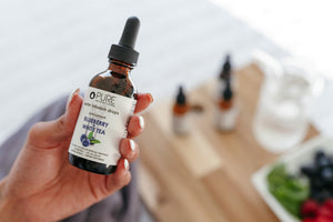 Hand holding a bottle of Super Fruit Extracts Water Infusion Drops - Blueberry + White Tea | Pure Inventions with blurred background featuring fruit and bottles.