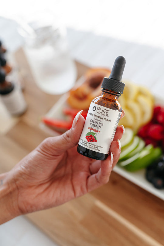 Hand holding a bottle of Antioxidant Green Tea Extract Water Infusion Drops - Raspberry | Pure Inventions with fresh fruit slices blurred in the background, promoting wellness supplements.