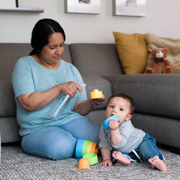 Woman sitting on the floor with a baby playing with colorful stacking cups next to a couch, holding a spray bottle from the 12oz Glass Bottle Cleaning Starter Kit | Force of Nature.