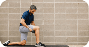 Man using RAD Muscle Flushing Kit | RAD for muscle recovery on thigh while kneeling on a mat against a gray brick wall, promoting wellness and body care.