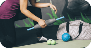 Person using the RAD Muscle Flushing Kit | RAD on their leg while kneeling on a yoga mat, with massage balls and a gym bag nearby, promoting muscle recovery and wellness.