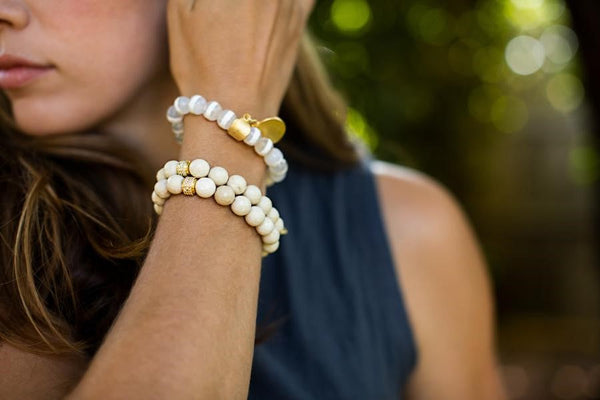 Close-up of a woman wearing the Natural Fossil Stone Beaded Stretch Bracelet | Meghan Bo, featuring white fossil stone beads and gold accents on her wrist against a blurred outdoor background.