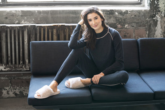 Young woman sitting on a dark sofa wearing cozy clothing and soft Anywhere Berber Slide | Lemon slippers, relaxing in a cozy living space with natural light.