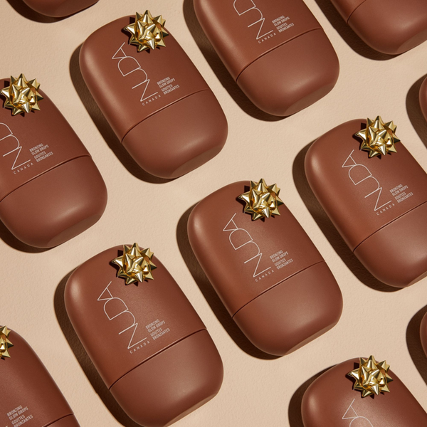 Multiple brown bottles of Bronzing Glow Drops | NUDA with gold decorative bows on a beige background, showcasing skincare product packaging for a bronzing glow effect.