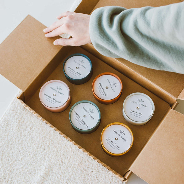 A person's hand opening a cardboard box containing six colorful glass jar soy candles, including Oakmoss + Amber Dignity Series Glass Jar Soy Candle | Calyan Wax Co. - 5.3 oz.
