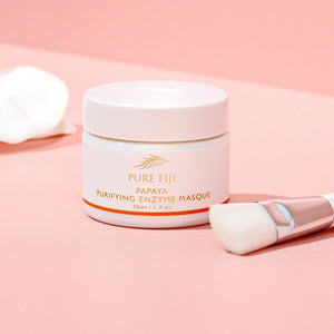 A jar of Papaya Purifying Enzyme Masque | Pure Fiji on a pink surface, accompanied by a soft cosmetic brush and a white cloth, highlighting skincare and wellness.