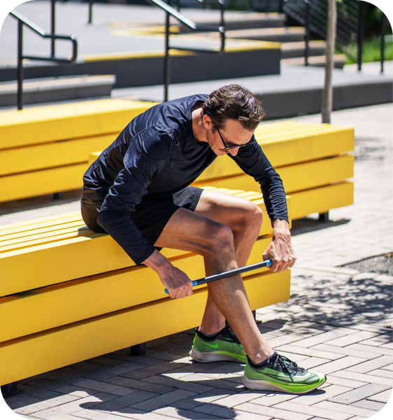 Man using RAD ROD | RAD muscle recovery tool on his calf while seated on a bright yellow bench outdoors in athletic wear.